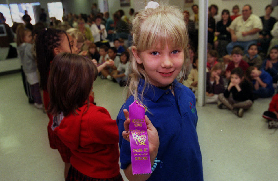 Young girl holding a “Music Award” ribbon in a school setting, surrounded by other children and adults in the background