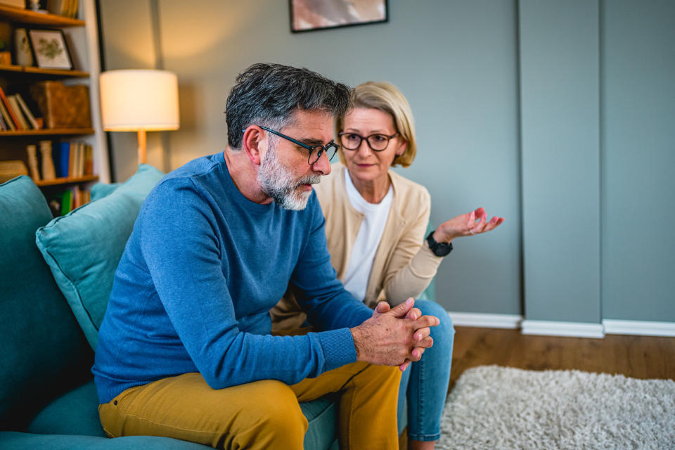 A man and a woman sit on a couch in a living room, engaged in a serious conversation. The man looks thoughtful, while the woman gestures as she speaks