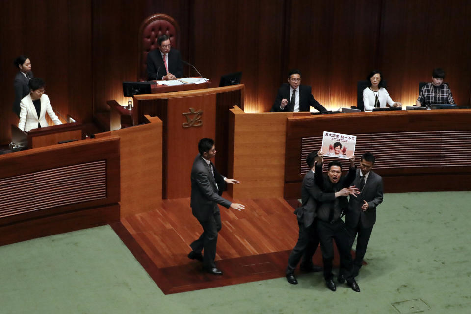 A pro-democracy lawmaker holds a plate card with picture of Hong Kong Chief Executive Carrie Lam with bloody hand on her face and slogan " Five Demands, No One Less" is forcibly removed from the chamber of the Legislative Council as Carrie Lam, left, delivers her speech at a question and answer session with lawmakers in Hong Kong, Thursday, Oct. 17, 2019. (AP Photo/Mark Schiefelbein)