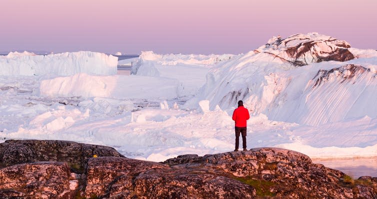 A scientist in red jacket stands before a glacier in Greenland.