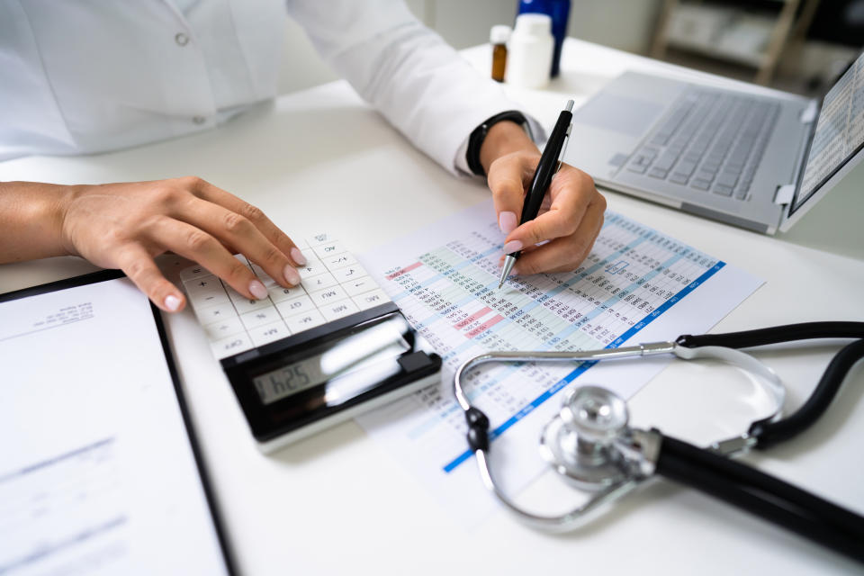 Close-up of a person using a calculator and writing on a desk with a stethoscope, laptop and calendar