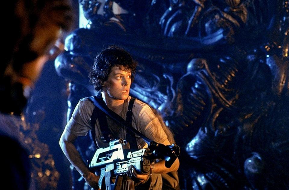 Sigourney Weaver with a large futuristic gun walks through a strange hallway