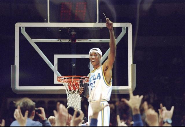 ed o'bannon smiling next to a basketball hoop and holding his arm in the air as others cheer in front of him and take his photo