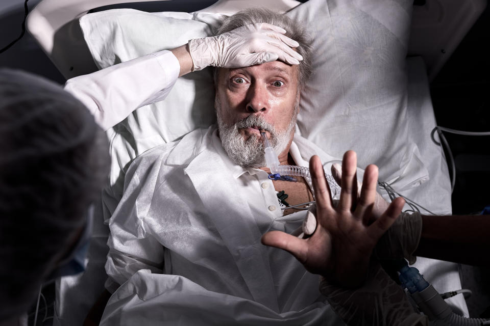 An older man with a surprised expression lies in a hospital bed, wearing a medical gown and connected to a ventilator. A medical professional's gloved hands are on his forehead and near his arm