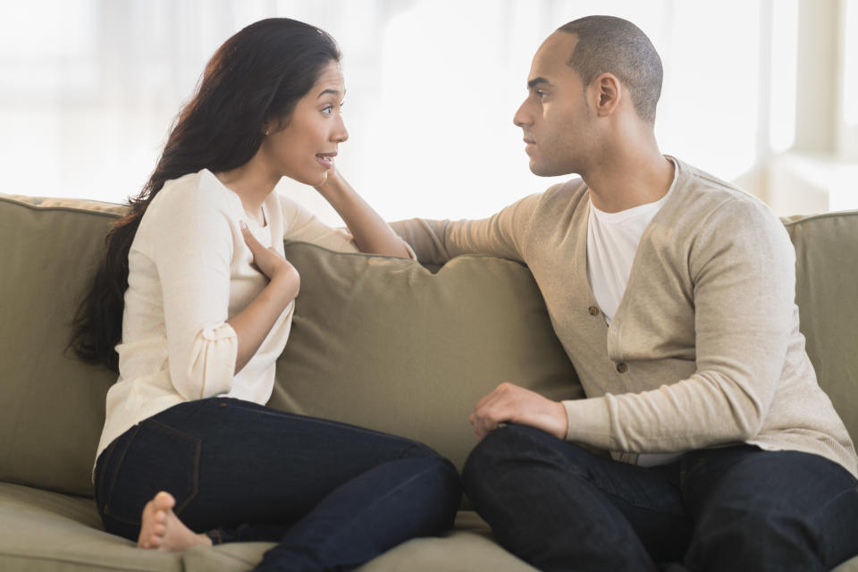 A couple sits on a sofa engaged in a serious conversation. The woman gestures with her hands while the man listens intently