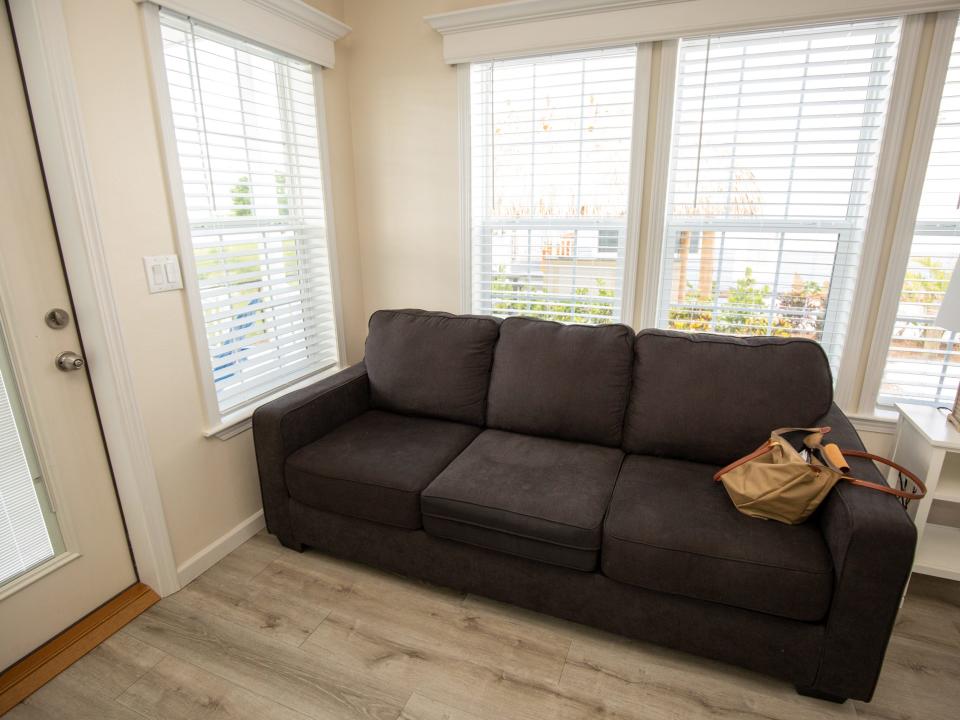 A couch with a purse in front of a wall of windows with blinds.