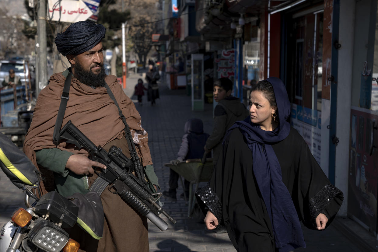 On a sidewalk, a woman glares at someone holding a military-style rifle.