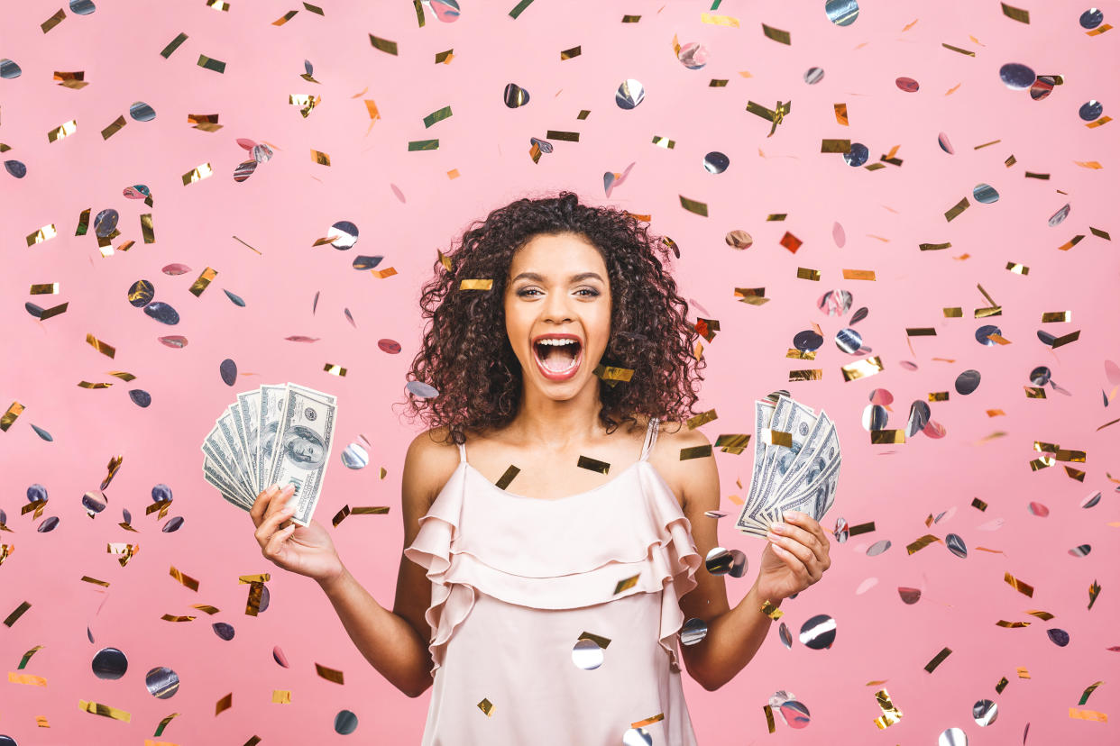 Black afro american girl won money. Happy young woman holding dollar currency satisfied isolated over pink background with confetti.