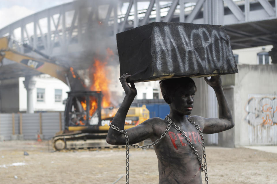 <p>A chained activist, with his body painted in black, lifts a fake stone bearing the name of French President Emmanuel Macron and the word “people” on his torso as he poses for photographers while a digger set ablaze by demonstrators is seen on background during the traditional May Day rally in the center of Paris, France, Tuesday, May 1, 2018. Each year, people around the world take to the streets to mark International Workers’ Day, or May Day. (Photo: Francois Mori/AP) </p>