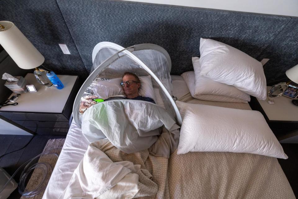 A man reads a book while lying in bed with his head encased in a plastic tent.