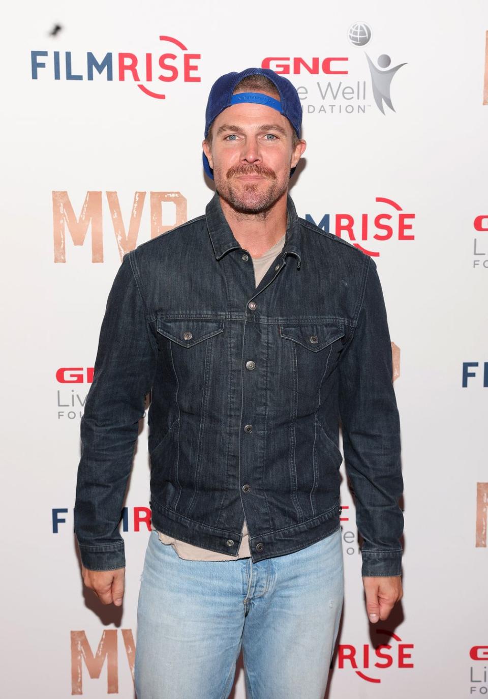 stephen amell stands looking at the camera with a neutral facial expression, he has brown hair and beard, he wears a cap backwards, grey tshirt, blue denim jacket and jeans