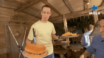 A man in a yellow shirt, carrying a large burger with a knife and a plate of nachos, serves food at an outdoor restaurant. The MOLA logo is in the top-right corner.