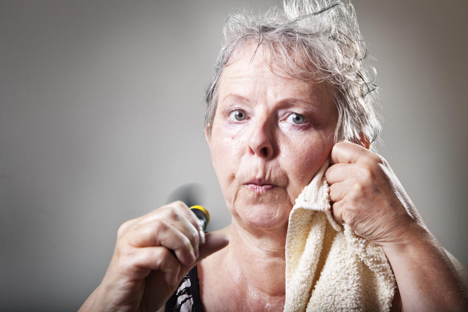 Woman in her fifties sweating, having hot flashes and trying to cool down with a small fan, drying face with towel