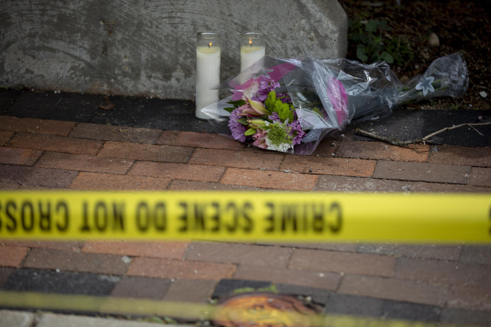 Two candles are lit on the sidewalk next to some flowers behind a yellow crime tape.