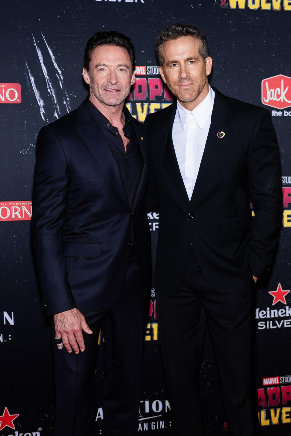 Hugh Jackman and Ryan Reynolds pose in suits at the "Deadpool 3" event. Hugh wears a dark suit, while Ryan dons a black suit with a white shirt