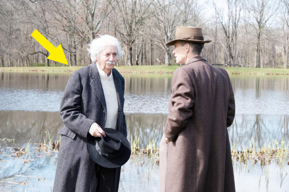 Albert Einstein and J. Robert Oppenheimer in conversation by a lake, both in period-appropriate overcoats and hats, with bare trees in the background