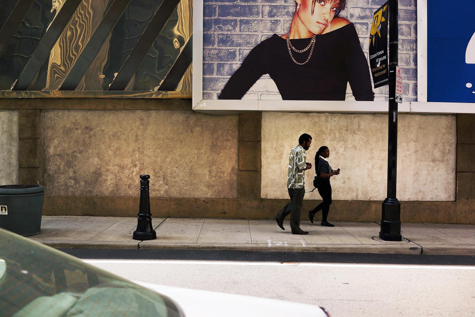 Pedestrians walk down a street in downtown NewarK, under a poster showing a glamorous young Black woman.