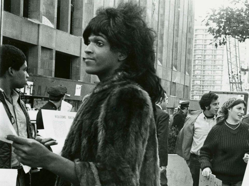 Black and white photo of Marsha P. Johnson in a fur coat handing out flyers in a busy street.