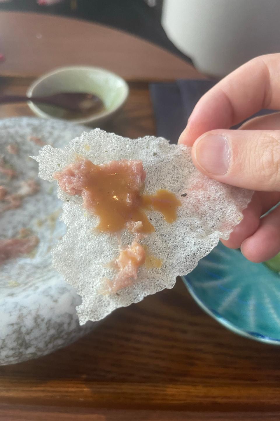 A hand holding a piece of rice cracker with a topping of raw tartare and sauce, with a marble bowl and other dishes visible in the background