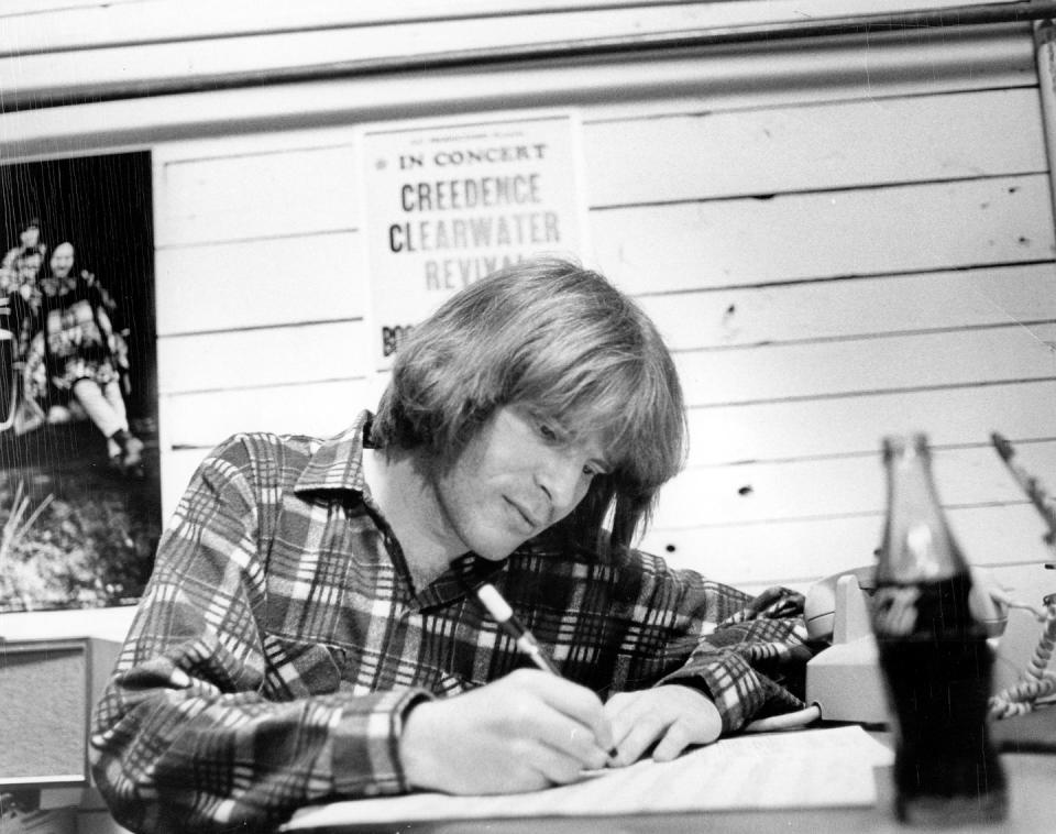 john fogerty sits at a desk and writes in this black and white photo, he is wearing a plaid shirt, next to him is a telephone, and a creedence clearwater revival poster hangs on the wall behind him