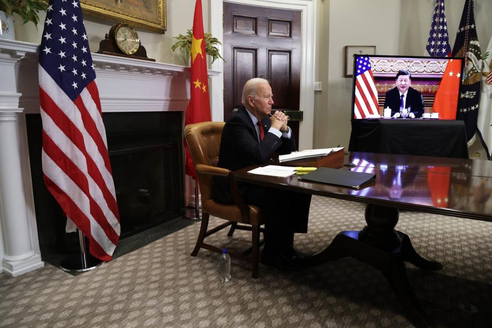 A seated man sits at. desk while another man is seen on a TV screen.