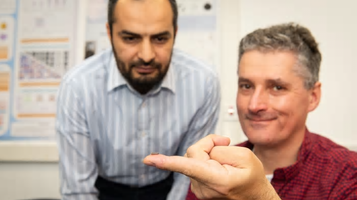 Alain Nogaret, en primer plano, mostrando una neurona artificial sobre la punta de su dedo. (Crédito imagen: Universidad de Bath).