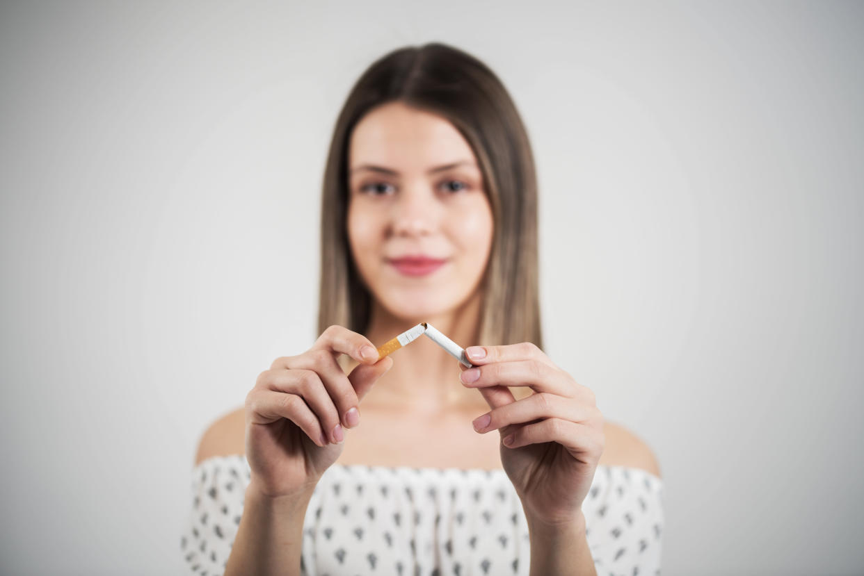 Close up portrait of young attractive woman breaking down cigarette to pieces. Studio shot selective focus isolated on grey. Addiction concept