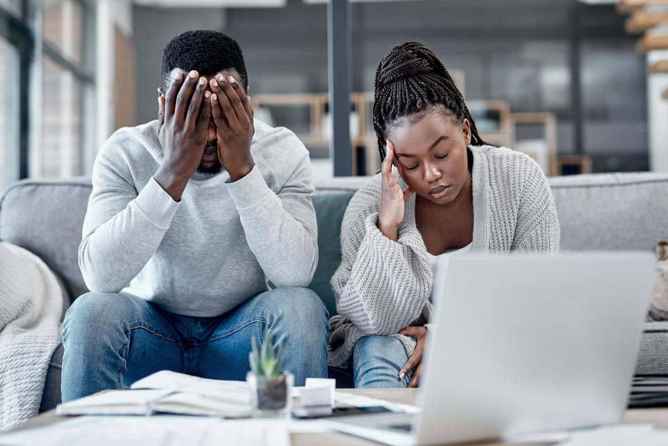 An African American couple sit on a couch looking exhausted, in front of an open laptop.