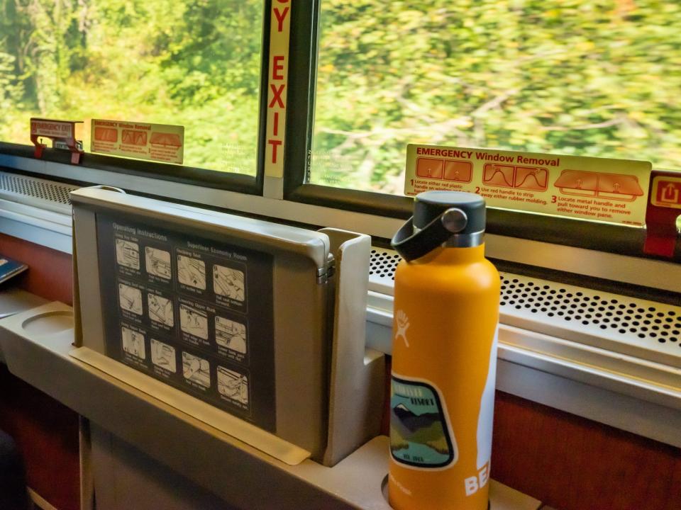 View of trees outside of window with orange water bottle on ledge by window inside Amtrak sleeper car