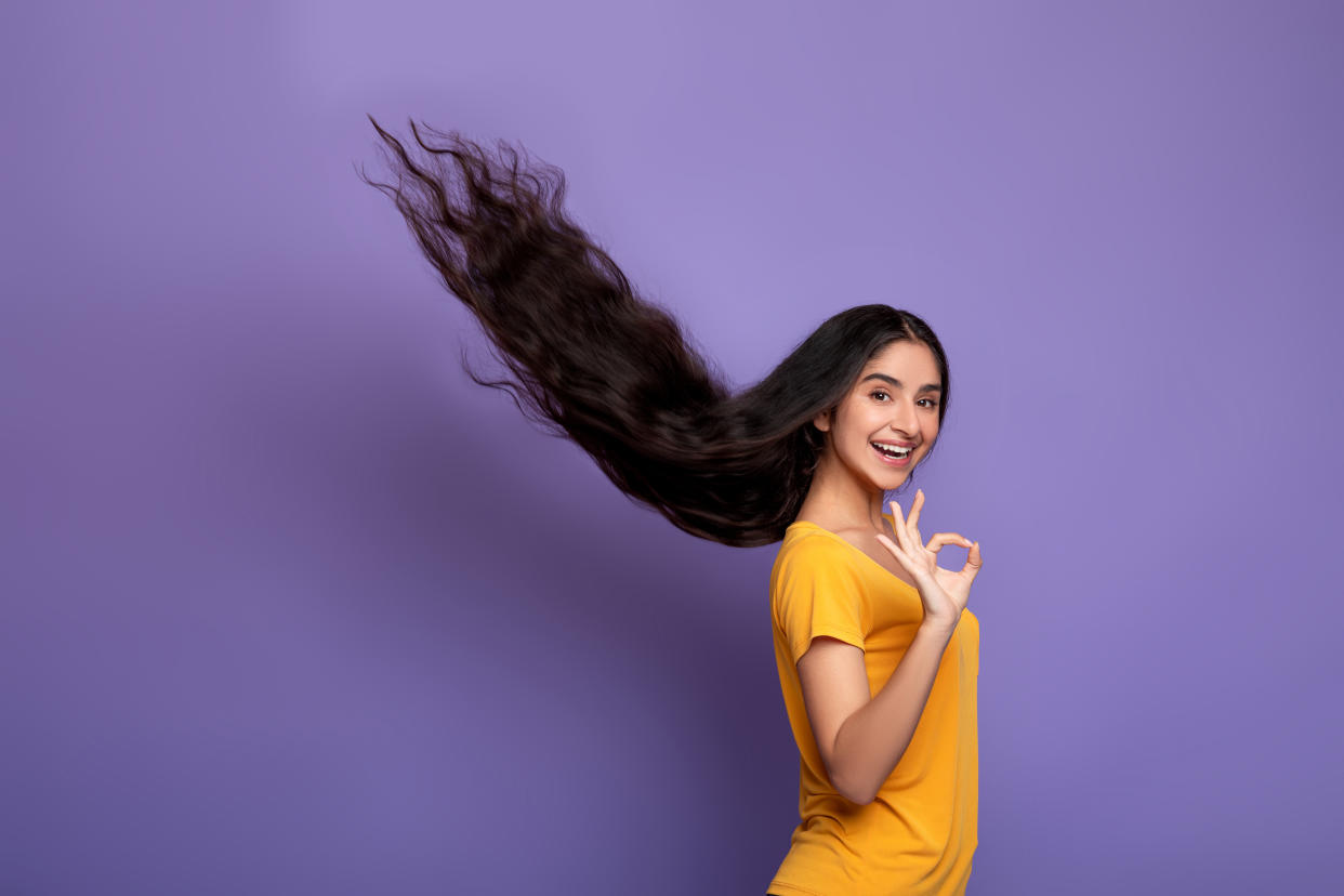 Hair Care Concept. Portrait of smiling indian model woman posing with long flying hair and showing okay sign gesture. Beautiful young lady standing isolated over purple studio background, copy space