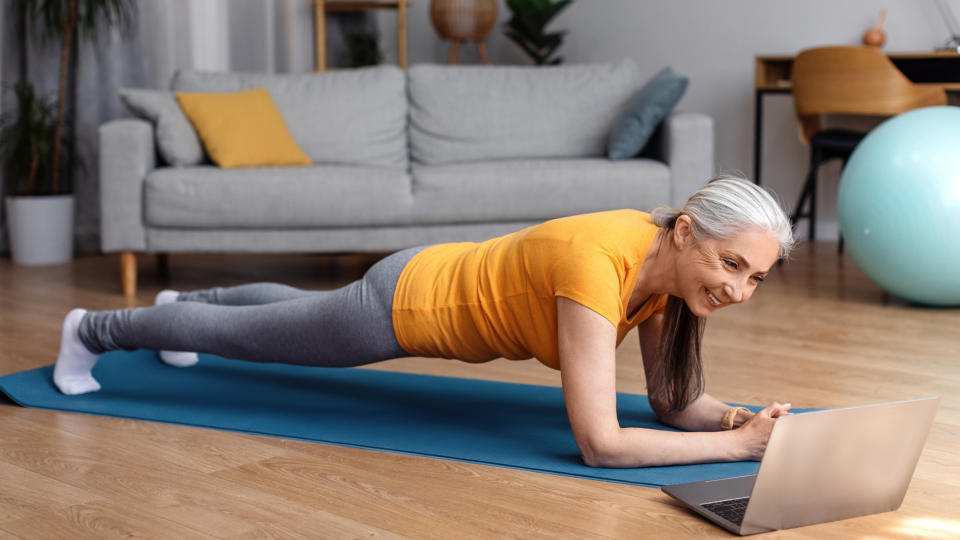 A woman in workout clothing performs a plank exercise, on a yoga mat in her living room. A laptop is open in front of her and we can see an exercise ball in the background
