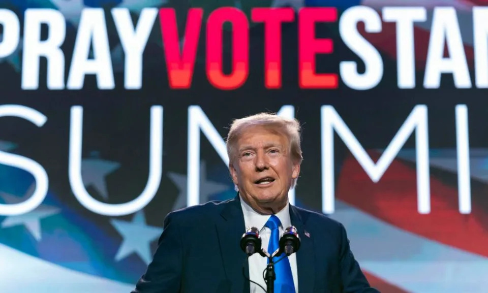 An older white man with fluffy reddish hair, a dark blazer, white shirt, and bright blue tie, stands at a lectern in front of a full-stage screen we can see says ‘Pray Vote Stand Summit’ against the backdrop of an American flag.