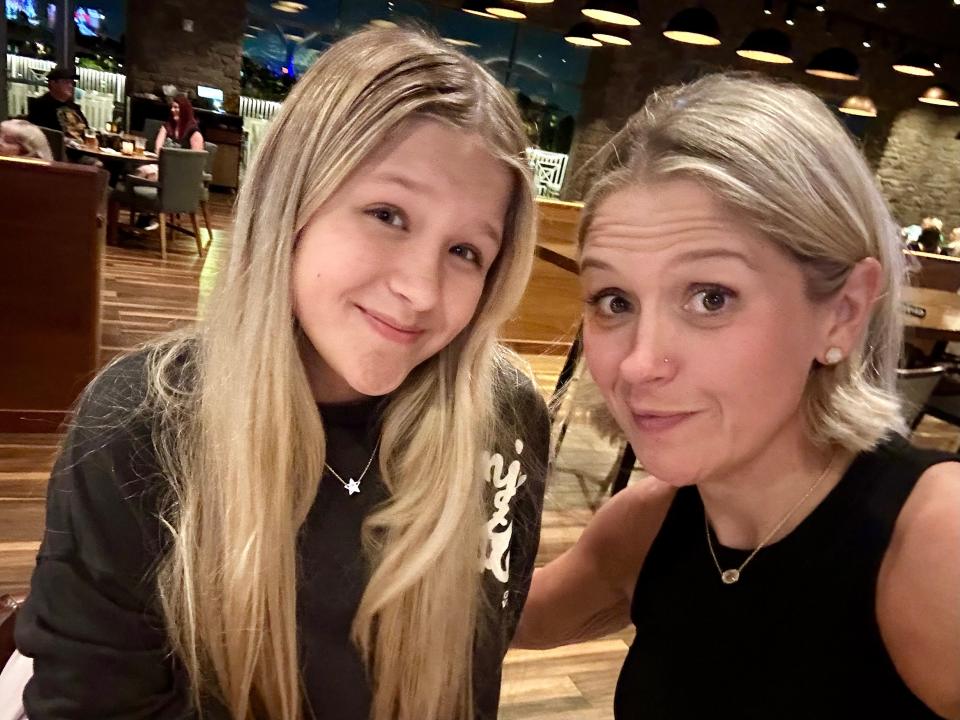 Terri and her daughter take a selfie in a restaurant. They smile slightly. Terri's daughter wears a black long-sleeved shirt and silver necklace with a star on it. Terri wears a black tank top and gold chain necklace with a light colored stone.