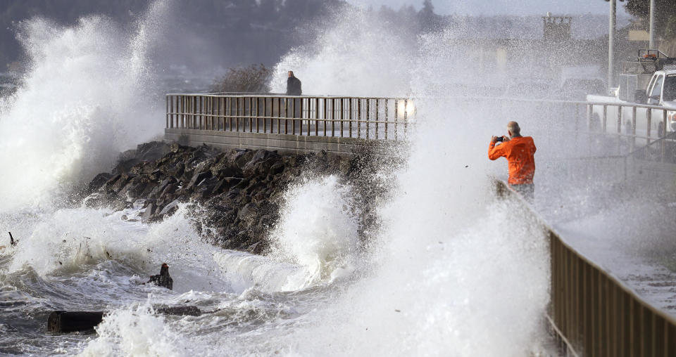 One man takes photos as another pedestrian looks out at waves crashing against a seawall at high tide during a windstorm Thursday, Dec. 20, 2018, in Seattle. More than 140,000 households and businesses lost power Thursday as strong winds toppled trees, closed roads and even trapped a trampoline between power lines in western Washington. A high wind warning remained in effect for much of area into Thursday evening, with gusts up to 60 miles an hour recorded earlier in the day. (AP Photo/Elaine Thompson)