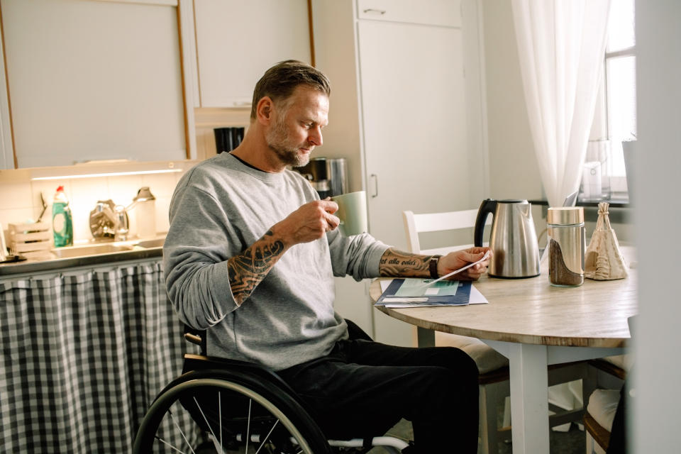 A man with tattoos, seated in a wheelchair, is holding a cup and reading papers in a kitchen