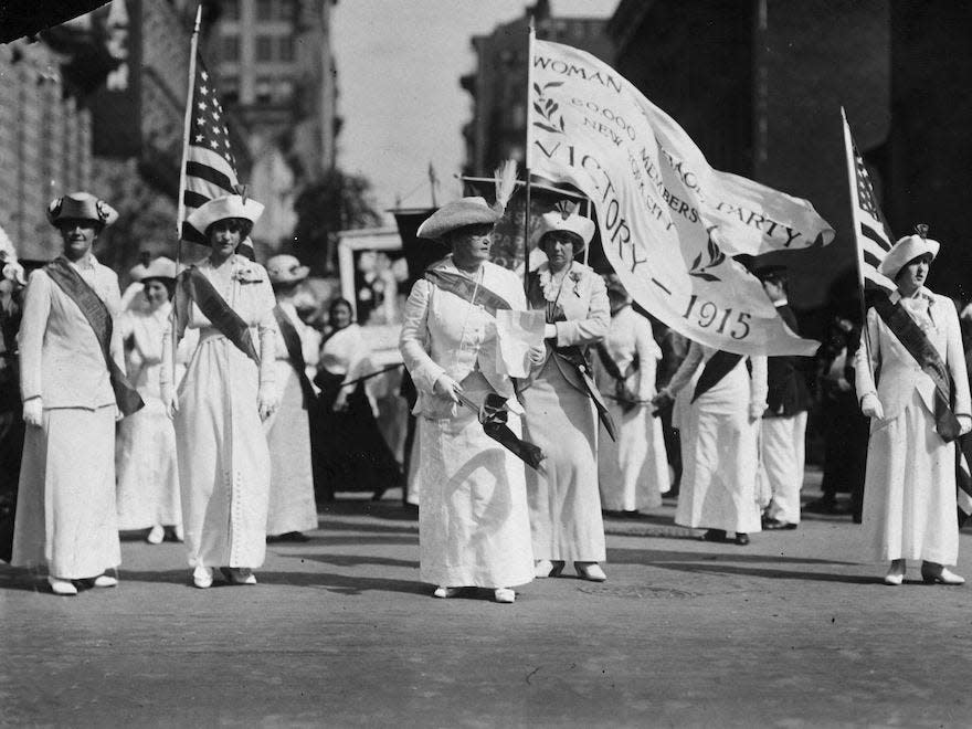 Suffragettes walking in a march wearing white dresses and holding flags.