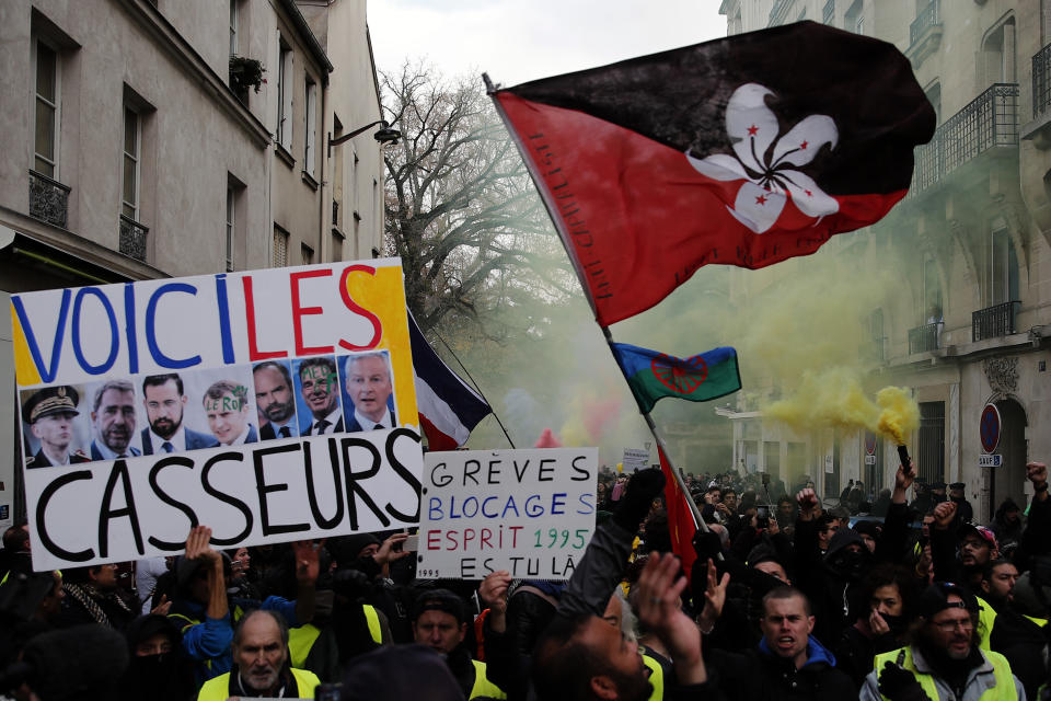A sign depicting French politicians and reading "Here are the troublemakers" and flags with flares are displayed during the yellow vests 56th round demonstration in Paris, Saturday, Dec. 7, 2019. A few thousand yellow vest protesters marched Saturday from the Finance Ministry building on the Seine River through southeast Paris, pushing their year-old demands for economic justice and adding the retirement reform to their list of grievances. (AP Photo/Francois Mori)