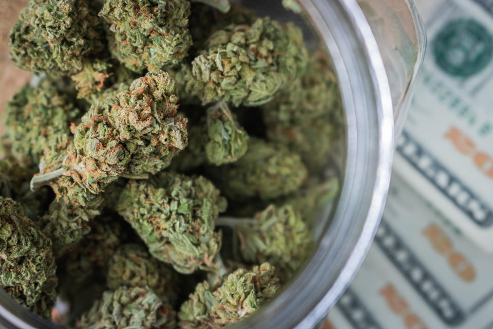 A jar filled with cannabis buds that's sitting atop a fanned pile of twenty dollar bills.