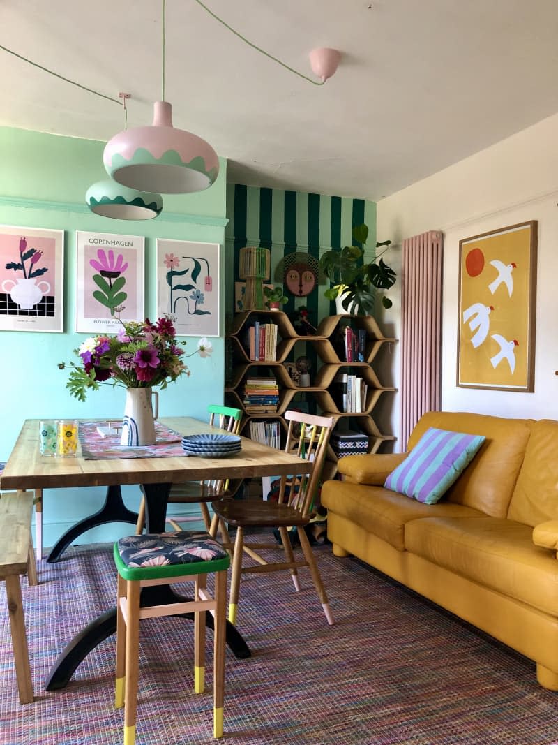 yellow leather couch, mint green wall, mint and dark green striped wall,  pink and green rounded lighting fixtures, hexagon shelves,  graphic art, plants,  blue and purple striped throw pillow, wooden bench, wooden chairs with green accents, pink and blue heathered rug