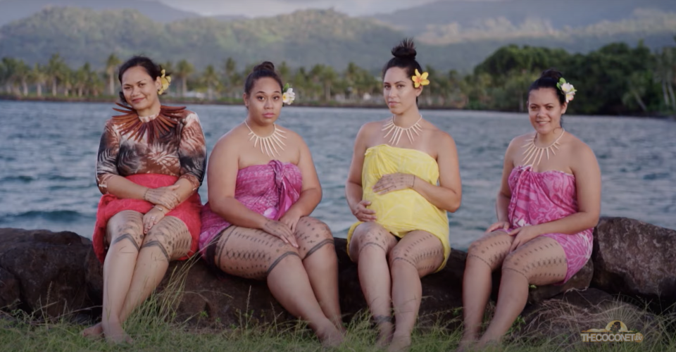 Four women in traditional Polynesian attire sitting on a rock with a tropical backdrop