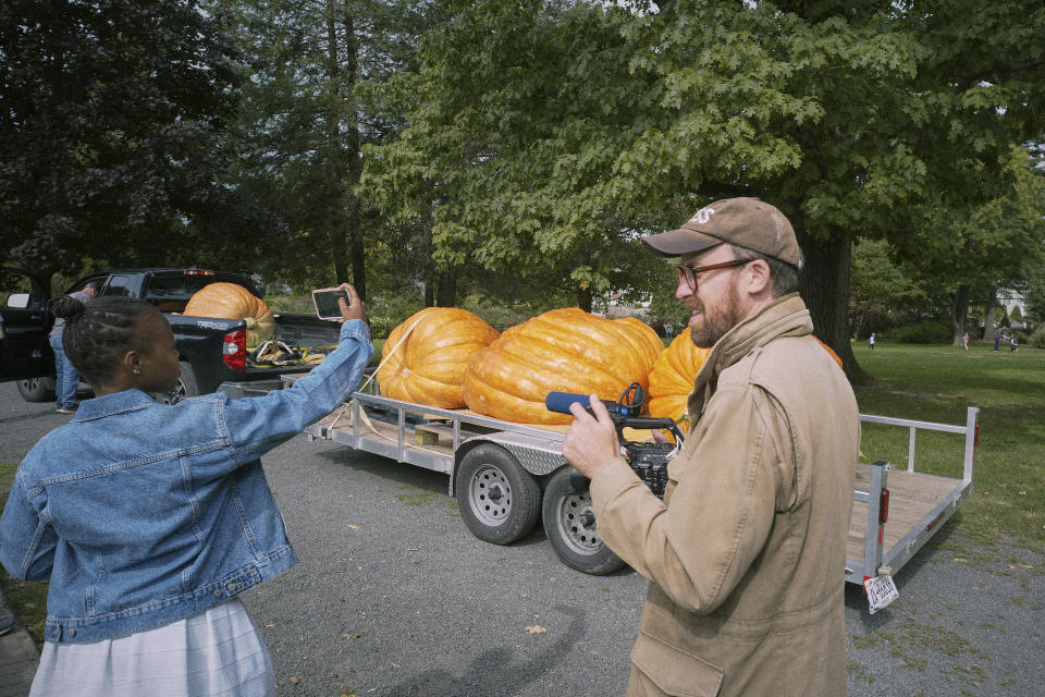 John Wilson stands next to a young girl in a jean jacket. Wilson is in profile holding a film camera and the girl is faced away from the camera, taking a picture of a trailer full of large pumpkins in the background.  