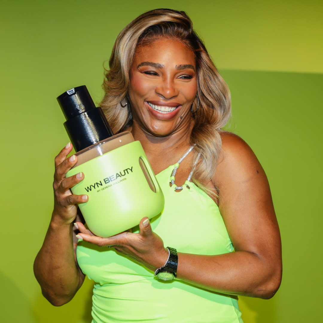  Serena Williams stands in front of a green wall holding a large bottle of wyn beauty foundation. 