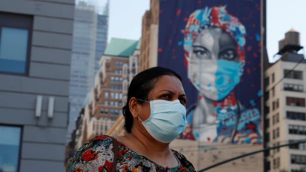 A woman wearing a mask stands in front of a New York building with a mural of a healthcare worker on it.