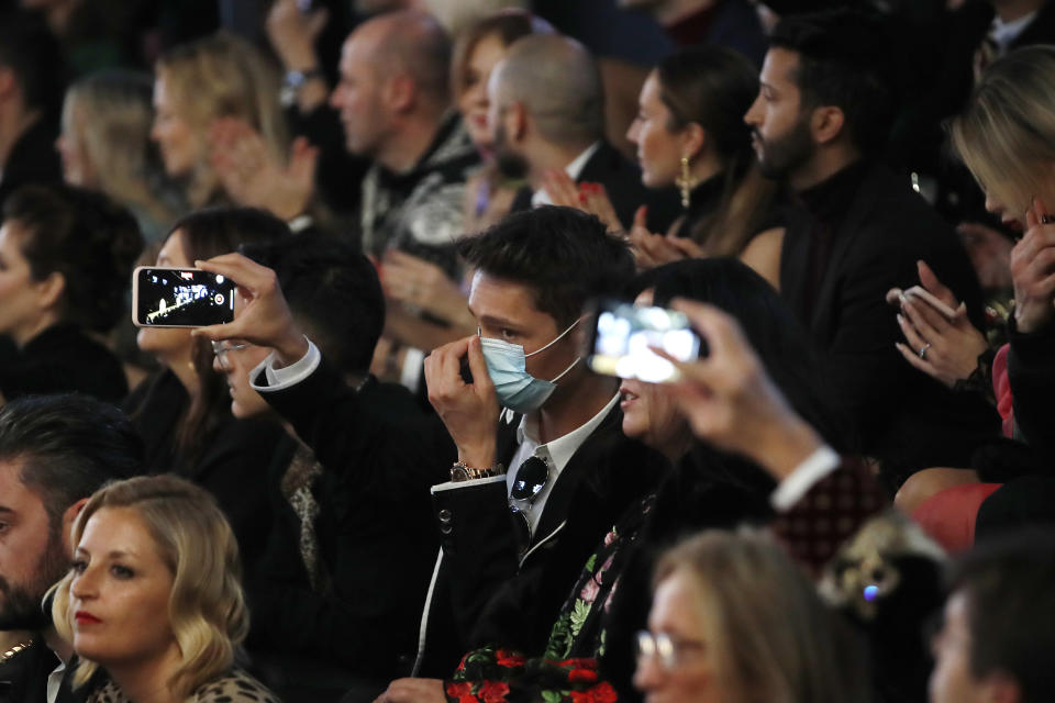 A man wearing a sanitary mask takes photos during the Dolce & Gabbana women's Fall Winter 2020-21 show, in Milan, Italy, Sunday, Feb. 23, 2020. After Giorgio Armani's last-minute decision to show his latest collection in an empty theater due to concerns about the new virus, the rest of Milan's runway shows scheduled for Sunday are to go ahead as planned, fashion officials confirmed. (AP Photo/Antonio Calanni)