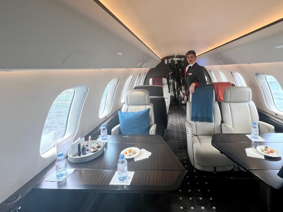 A flight attendant stands in the main seating/dining area on board a VistaJet Bombardier Global 7500 at Farnborough Airport, where nine cream leather seats are visible.