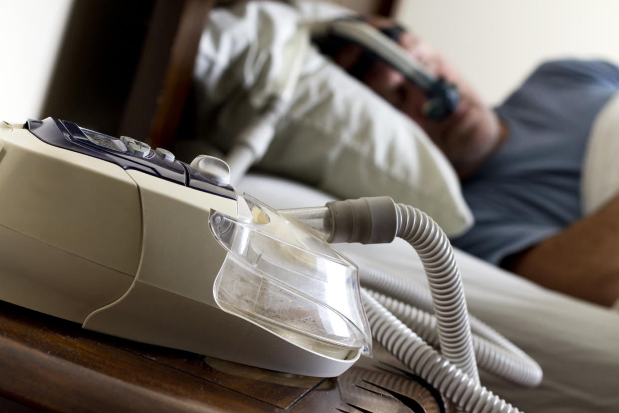 A man sleeps, wearing a CPAP mask, with tubes and the CPAP machine in the foreground.