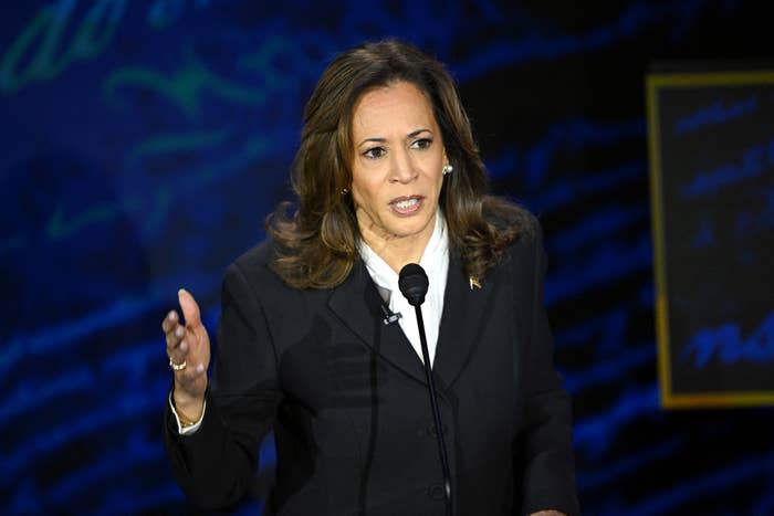 Kamala Harris speaks passionately at a podium during a public event. She wears a formal, dark blazer over a blouse