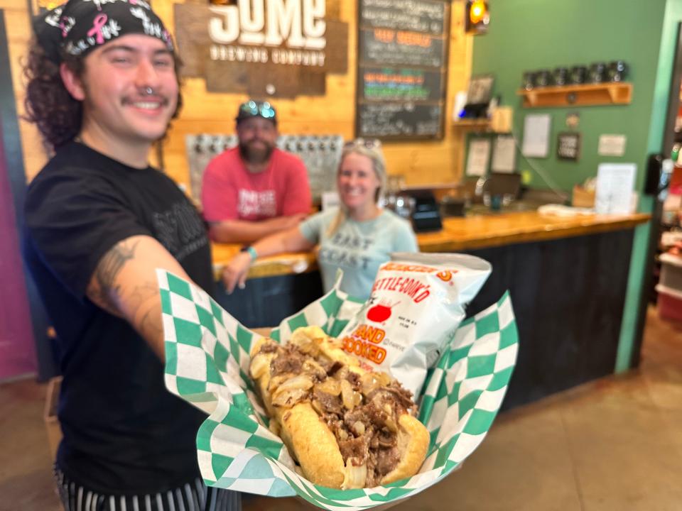 Chef Cam Pappas holds a Philly cheesesteak that will soon be served from the recently added menu at SoMe Brewing Company. Also pictured are co-owner David Rowland and his sister, General Manager Sarah Rowland.