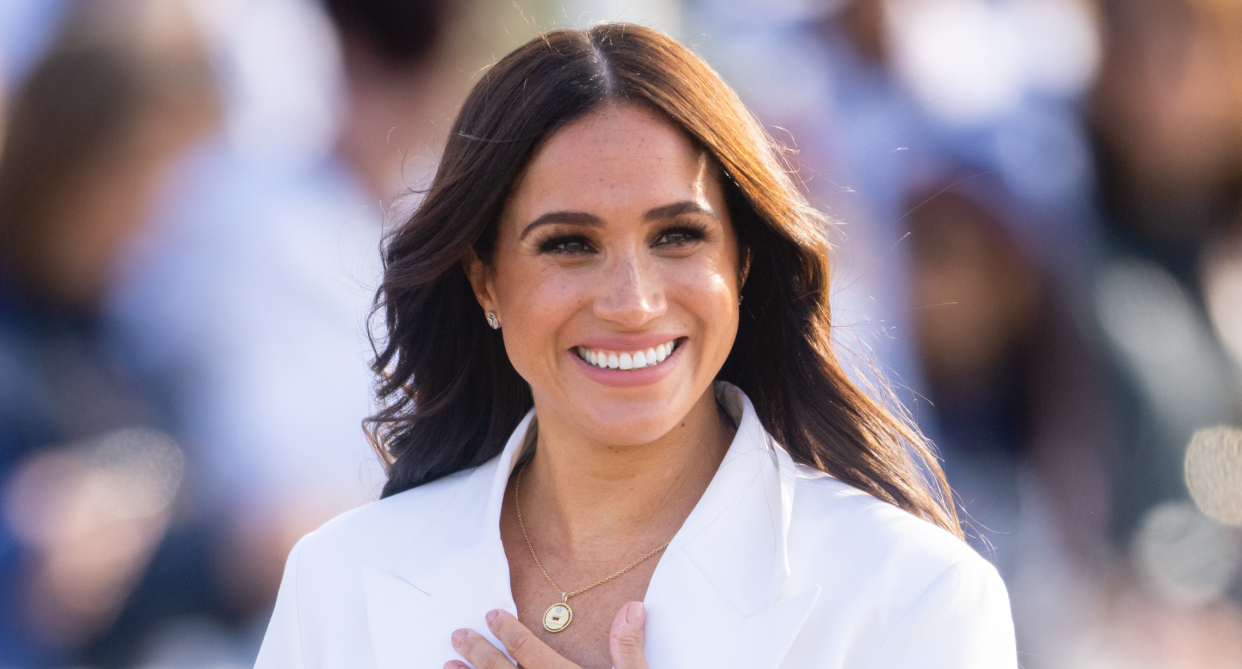 meghan markle with hair down smiling wearing white blazer and gold pendant necklace with hand on chest