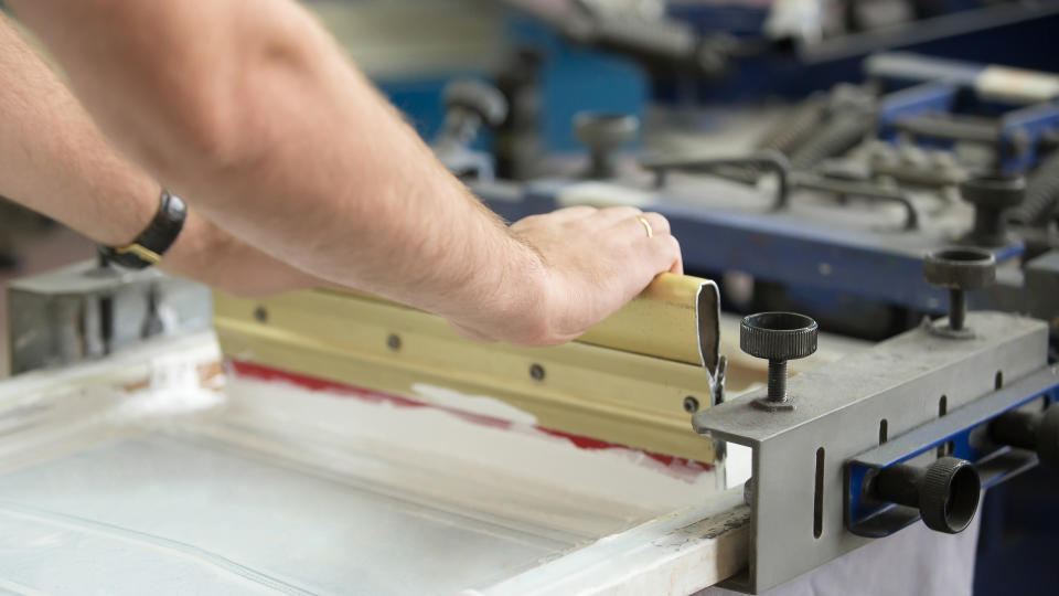 Close up of male hands exploring traditional squeegee, creating an image on white fabric clothing by pressing ink through a screen with areas blocked off by a stencil.